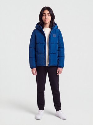 Jeune personne debout, portant une doudoune à capuche bleue, une chemise blanche, un pantalon noir et des baskets blanches, sur un fond clair uni.