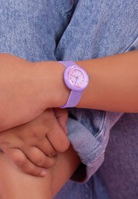Montre en silicone violet avec un boîtier rond, un cadran rose et blanc, et des chiffres audacieux. Le bracelet présente un motif scintillant, reposant sur une main.