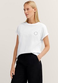 Weißes Baumwoll-T-Shirt mit umgeschlagenen Kurzärmeln und einem kleinen runden schwarzen Grafikdruck auf der Brust. Kombiniert mit schwarzen Hosen.