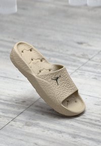 Μπεζ σανδάλι slide με ανάγλυφο σχέδιο, ανοιχτό μπροστά και μαύρη λεπτομέρεια με λογότυπο. Κατασκευασμένο από υλικό καουτσούκ με υφή.