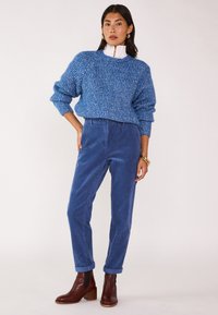 Maglione blu a maglia con una finitura testurizzata, abbinato a pantaloni in corduroy blu e stivali alla caviglia marroni. Gli accessori includono bracciali d'oro.