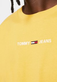 Yellow cotton t-shirt featuring white embroidered text "TOMMY" and "JEANS" with a small graphic logo of red, white, and blue stripes.