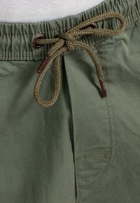 Pantalones cortos de algodón verde con cintura ajustable por cordón, presentando cordón trenzado y extremos de metal marrón, además de un bolsillo lateral y una textura suave.