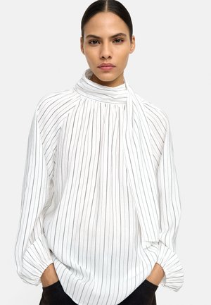 Kobieta w białej bluzce z czarnymi pionowymi paskami i wiązaniem przy szyi, stojąca z rękami w kieszeniach na jednolitym tle.