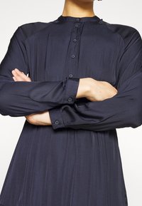 Personne portant une chemise à manches longues bleu marine foncé boutonnée, les bras croisés sur la poitrine, sur un fond clair uni.