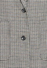Παλτό με σχέδιο καρώ τύπου houndstooth με δύο μαύρα κουμπιά και δύο τετράγωνες μπροστινές τσέπες σε μαύρους, λευκούς και μπεζ τόνους.