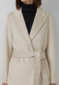 Beige Wolle Mantel mit taillierter Silhouette, ausgestattet mit einem Gürtel in der Taille, spitzen Revers und einer Tasche mit einer goldfarbenen Knopfakzent.