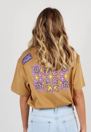 T-shirt surdimensionné marron avec un design floral en violet, rose et jaune au dos ; présente un petit logo sur l'épaule gauche. Matériel en coton.