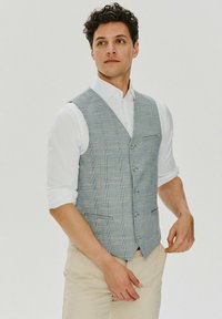 Szary kamizelka w kratę z niebieskimi akcentami, z dekoltem w serek, pięcioma guzikami i kieszeniami po bokach, noszona na białej koszuli i beżowych spodniach.