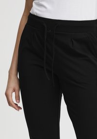 Černé joggingové kalhoty s elastickým pasem, nastavitelným šňůrkou a sklady. Vyrobeny z měkké, pružné tkaniny pro pohodlí.