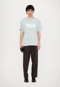 Ανοιχτό μπλε βαμβακερό μπλουζάκι με λευκή ριγέ εκτύπωση και αριθμό "6" στο μπροστινό μέρος, συνδυασμένο με σκούρες καφέ παντελόνι και μαύρα παπούτσια.