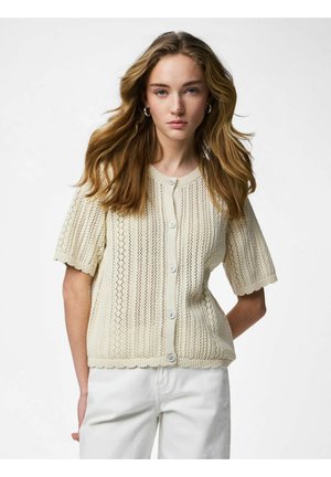 Junge Frau mit welligem blondem Haar, die eine cremefarbene kurzärmelige Strickjacke mit Knöpfen und weiße Hosen trägt, vor einem weißen Hintergrund stehend.