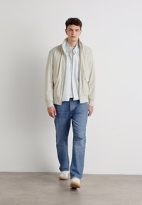 Lys beige bomberjakke med to sidelommer, båret over en hvit skjorte og blå jeans, sammen med hvite sneakers på en nøytral bakgrunn.