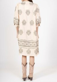 Robe rose clair avec une broderie florale grise complexe, des manches bouffantes et des poches latérales, associée à des bottes cowboy beiges.