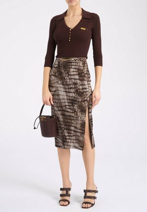 Femme portant un haut marron côtelé avec des boutons dorés, une jupe midi à motifs avec une fente sur le côté, un sac à main marron foncé et des sandales à talons marron.