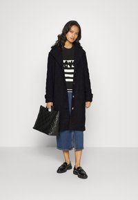 Lunga giacca nera con un motivo testurizzato, chiusura con bottoni, indossata sopra un maglione a righe, abbinata a una gonna di jeans e mocassini neri. Borsa a forma di tote nera trapuntata.