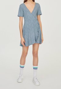 Modré zavinovací šaty s drobným bílým květinovým vzorem, výstřihem do V, krátkými rukávy a volánkovým lemem. V kombinaci s bílými teniskami a proužkovanými ponožkami.