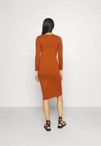 Rebrasti haljina srednje dužine u boji izgorene naranče s dugim rukavima i prilagodljivim siluetom, uparena s crnim sandalama na remen.