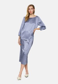 Satin two-piece outfit in light blue, featuring a long-sleeve top with a boat neckline and a knee-length skirt, worn with cream-colored shoes.