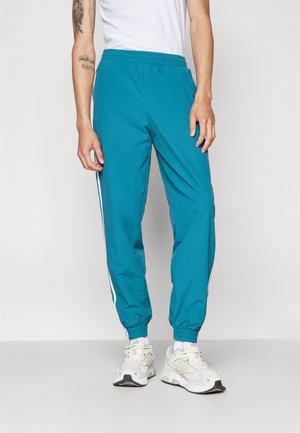 Persoon die teal joggingbroeken draagt met witte zijstrepen, witte sneakers en een wit mouwloos topje, staand tegen een neutrale achtergrond.