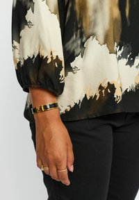Sort og beige mønstret bluse med trekvartærmer og elastiske manchetter. Et guld- og sort armbånd pryder håndleddet, suppleret med ringe.