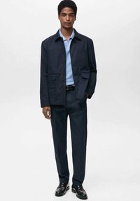 Giacca navy con chiusura a bottoni e tasche applicate, abbinata a una maglietta a maglia azzurra chiara e pantaloni scuri, completata da scarpe nere.