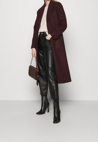 Femme portant un long manteau bordeaux, un top beige, un pantalon en cuir noir, des bottines à talons hauts noires, tenant un sac à main matelassé marron avec une chaîne dorée.