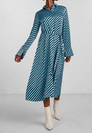 Femme portant une robe midi à manches longues rayée en diagonale bleu et noir avec une ceinture, associée à des bottines blanches.