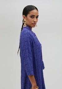 Lila tunika med texturerad tyg, med diamantmönster och trekvartsärmar. Anmärkningsvärd tofsdetalj och lager av pärlhalsband.