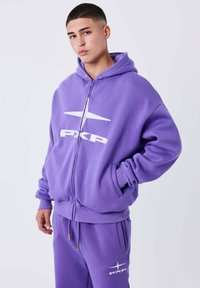 Sudadera con cremallera en color púrpura, con un logo blanco, capucha y bolsillos tipo canguro. El material parece suave y texturado, combinada con pantalones de chándal a juego.
