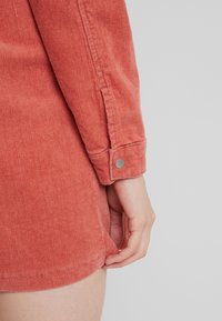 Kordurové šaty v rezavé barvě s detaily na manžetách. Tkanina má vroubkovanou texturu a lem, který se po stranách zakřivuje vzhůru.