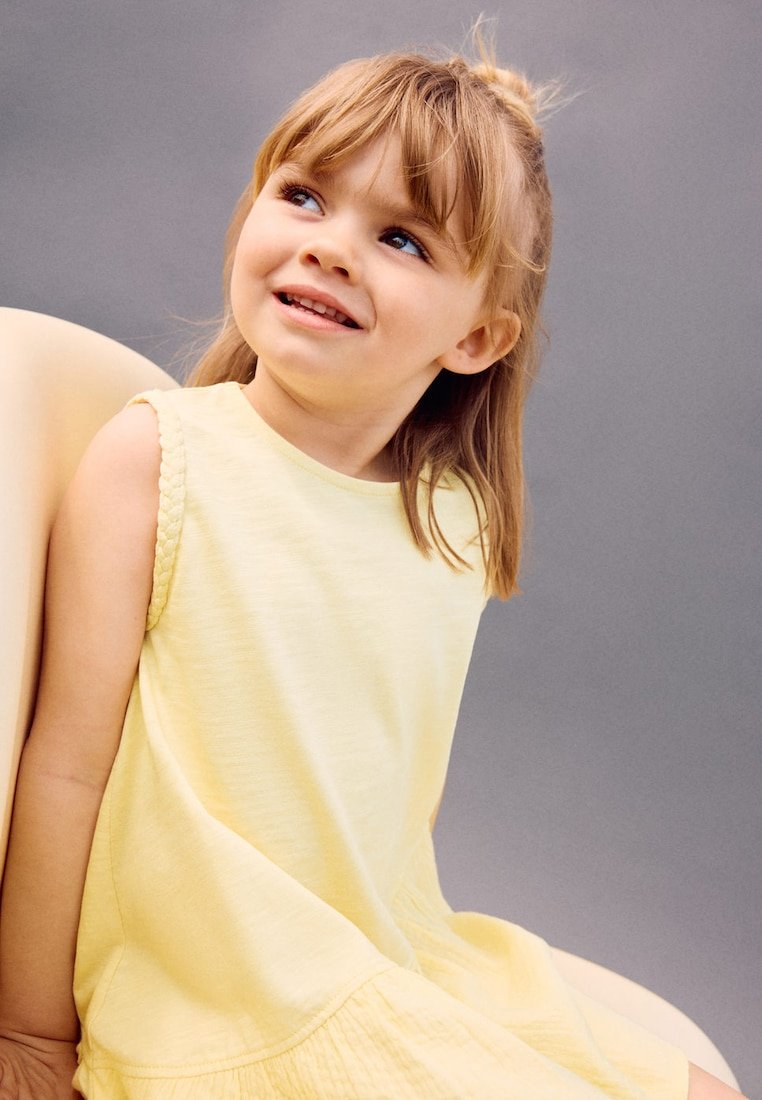 Jeune fille aux cheveux châtain clair portant une robe jaune sans manches, assise et souriante, regardant légèrement vers le haut sur un fond gris.