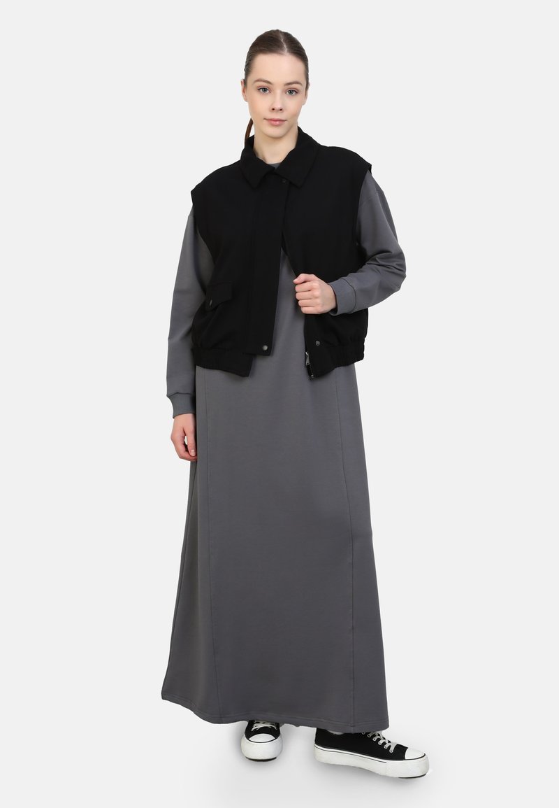 Schwarze ärmellose Jacke über einem langen grauen Kleid. Die Jacke hat einen Kragen und Knöpfe vorne, während das Kleid eine glatte Textur aufweist. Schwarze Sneakers runden den Look ab.