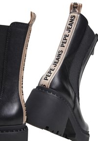 Botas negras hasta el tobillo con un tacón grueso, que cuentan con un panel de tela y una tira elástica marrón con el logo "PEPE JEANS". Superficie de cuero suave.