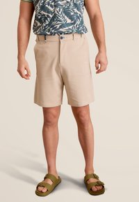 Beige korte broek met een rechte voorkant, riemlussen en zijzakken, gecombineerd met een gestreept blauw en groen shirt en olijfgroene sandalen.
