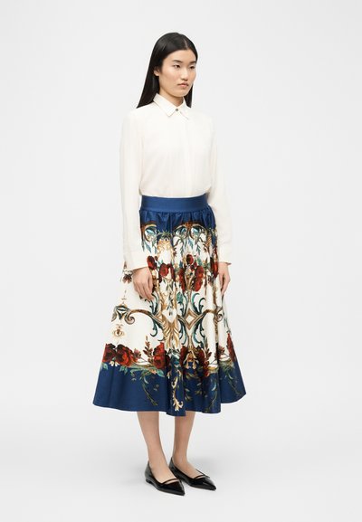 Femme debout portant un chemisier blanc à manches longues et une jupe taille haute, longueur cheville, avec des motifs bleus et floraux, ainsi que des ballerines noires.