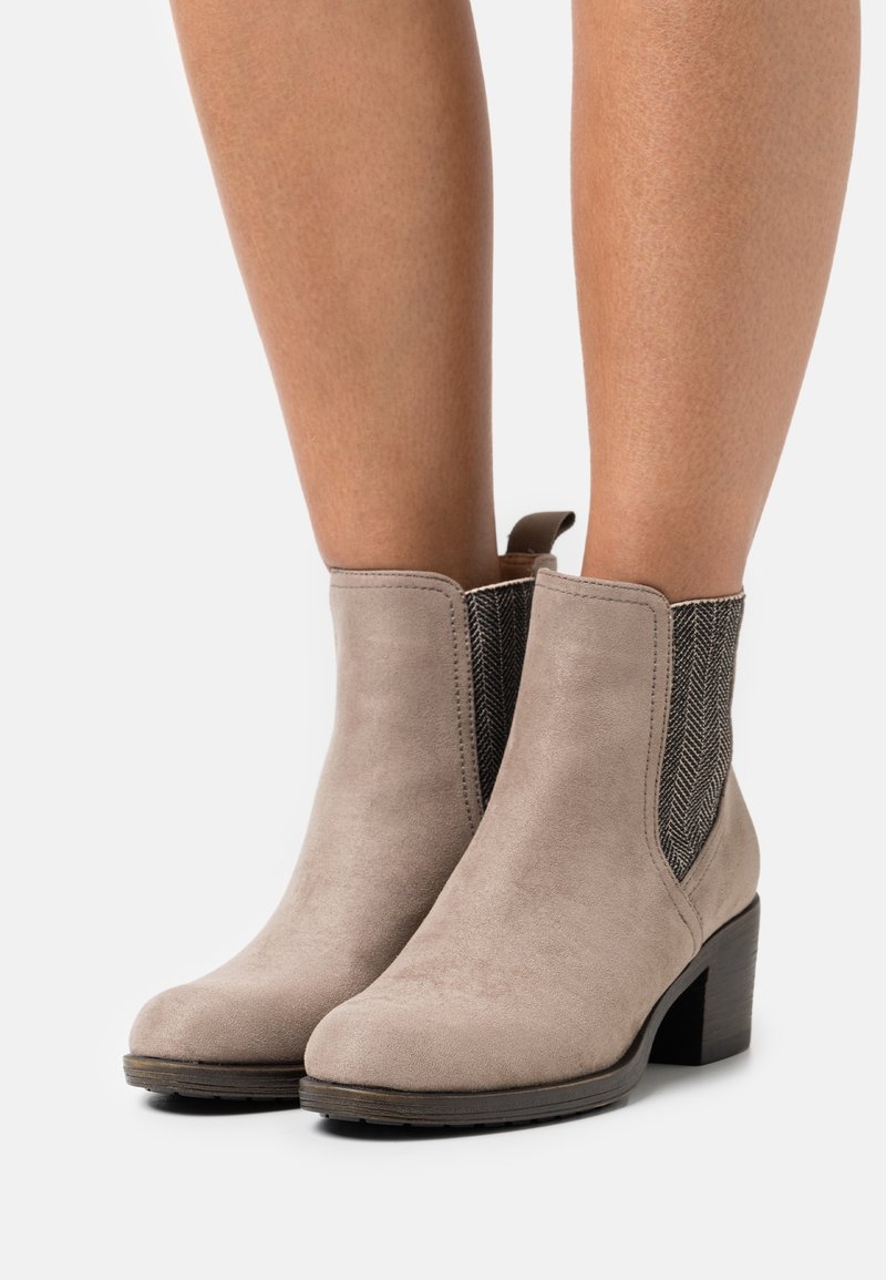 Bottes en daim taupe montant à la cheville avec des panneaux élastiques latéraux noirs en chevrons et des talons blocs portées sur des jambes nues.