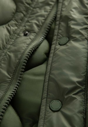 Lähikuva oliivinvihreästä tikatusta takista, jossa näkyvä vetoketju ja pyöreät napit, joissa on brändin nimi kaiverruksena.