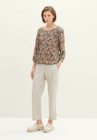 Bluse mit Blumendruck in Orange, Grün und Weiß mit 3/4-Ärmeln, kombiniert mit hellbeigen Cargo-Hosen und cremefarbenen Slippern. Flache Sohle.