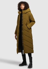 Olijfgroene gewatteerde lange jas met een capuchon, ritsluiting, diagonale stiksels en zijzakken, gecombineerd met zwarte laarzen en legging.