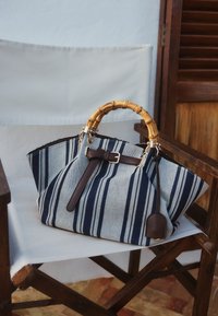 Sac à main en tissu rayé avec des anses en bambou et un détail de boucle en cuir marron reposant sur une chaise en bois avec une assise en toile blanche.