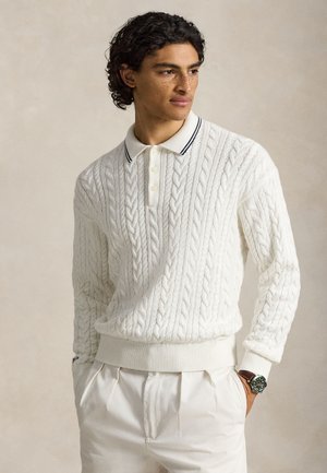 Ung mand med krøllet hår og fregner iført en hvid kabelstrikket sweater og matchende hvide bukser, hænderne i lommerne, ser mod venstre.