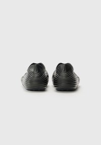 Black slip-on shoes with a textured, wavy design, featuring a rounded toe and low back. Made from flexible material, with a flat, durable sole.