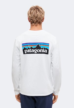 Person med kort blondt hår iført en hvid langærmet skjorte med et farverigt Patagonia-logo på ryggen, stående mod en ensfarvet baggrund.