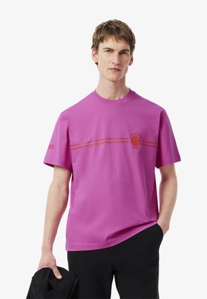 Rosa Baumwoll-T-Shirt mit Rundhalsausschnitt, kurzen Ärmeln und einem braunen Grafikdesign mit den Worten "LACOSTE PARIS". Glatte Textur.