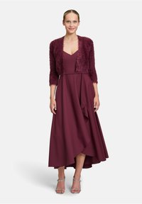 Robe bordeaux avec un corsage ajusté, une jupe évasée et un ourlet asymétrique, accompagnée d'un shrug en fausse fourrure texturé assorti. Sandales argentées à lanières.