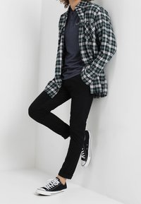 Rutig flanellskjorta i grön och vit, lager på lager över en marinblå piké. Svarta slim-fit-byxor och svarta sneakers fulländar looken.