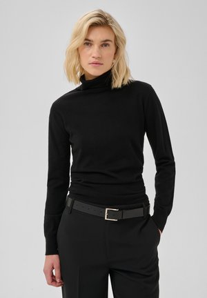 Femme aux cheveux blonds porte un pull à col roulé noir, un pantalon noir et une ceinture noire, debout avec une main dans la poche.