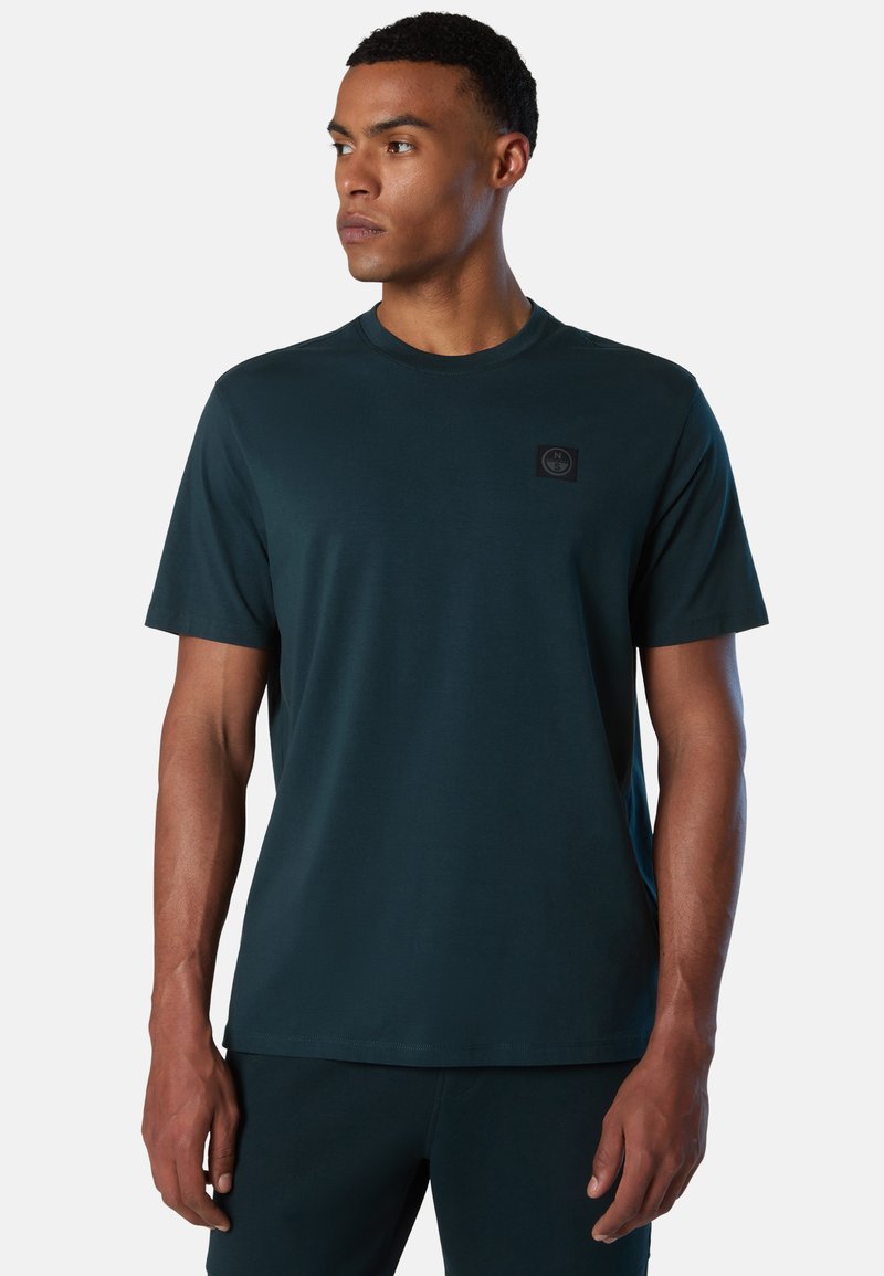 Hombre vistiendo una camiseta de manga corta verde oscuro con un pequeño logo en el pecho, mirando hacia su izquierda contra un fondo claro y liso.