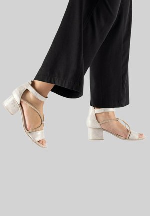 Pieds portant des sandales à talons blocs blanches avec des lanières, associées à un pantalon noir à jambes larges, sur un fond gris uni.